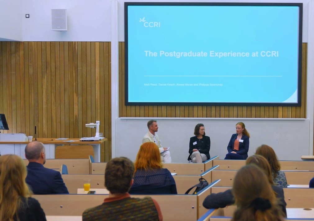 three people having a conversation in front of an audience with a screen behind them