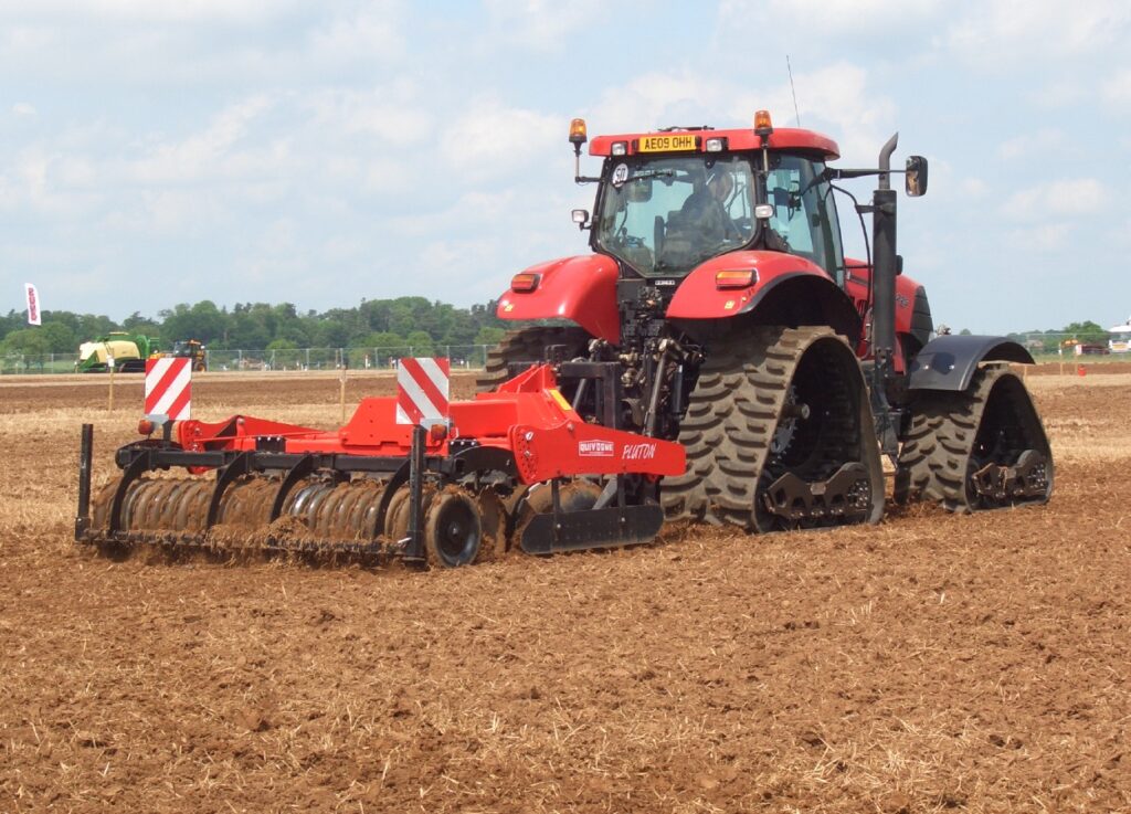 A large red tractor with tracks towing a red soil roller in a field of exposed soil