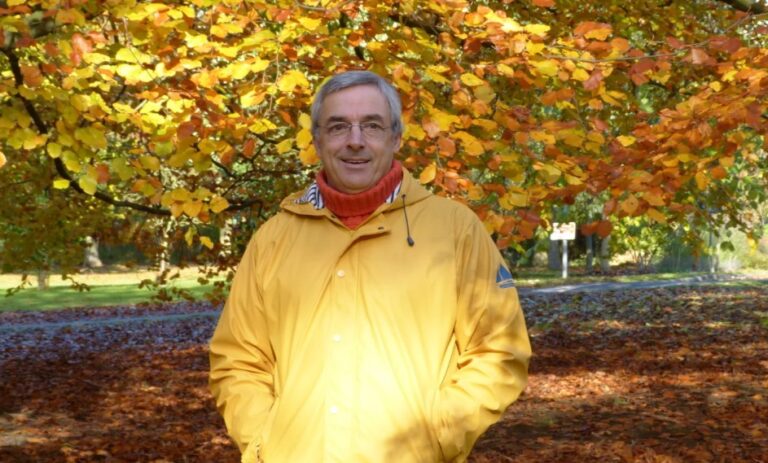 Professor Nigel Curry wearing a yellow coat. He is stood in front of an autumnal tree and gently smiling. He will be missed.